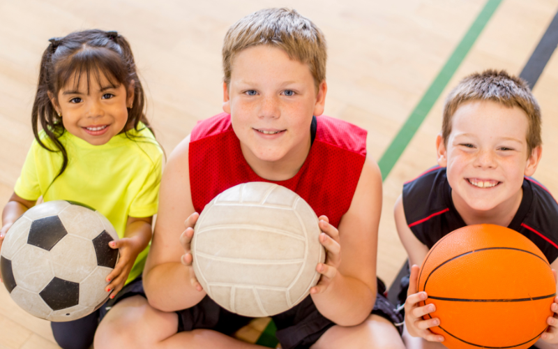 Three Children Are In A Sportscentre, They Are Smiling Holding Footballs And A Basketball