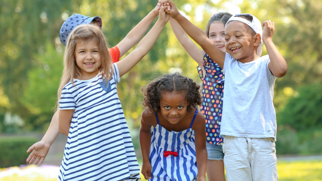 Five Children Are Playing Together Outside, Smiling And Laughing. Four Are Holding Hands To Create An Arch And The Fifth Child Is Crouched Down Running Through The Arch.