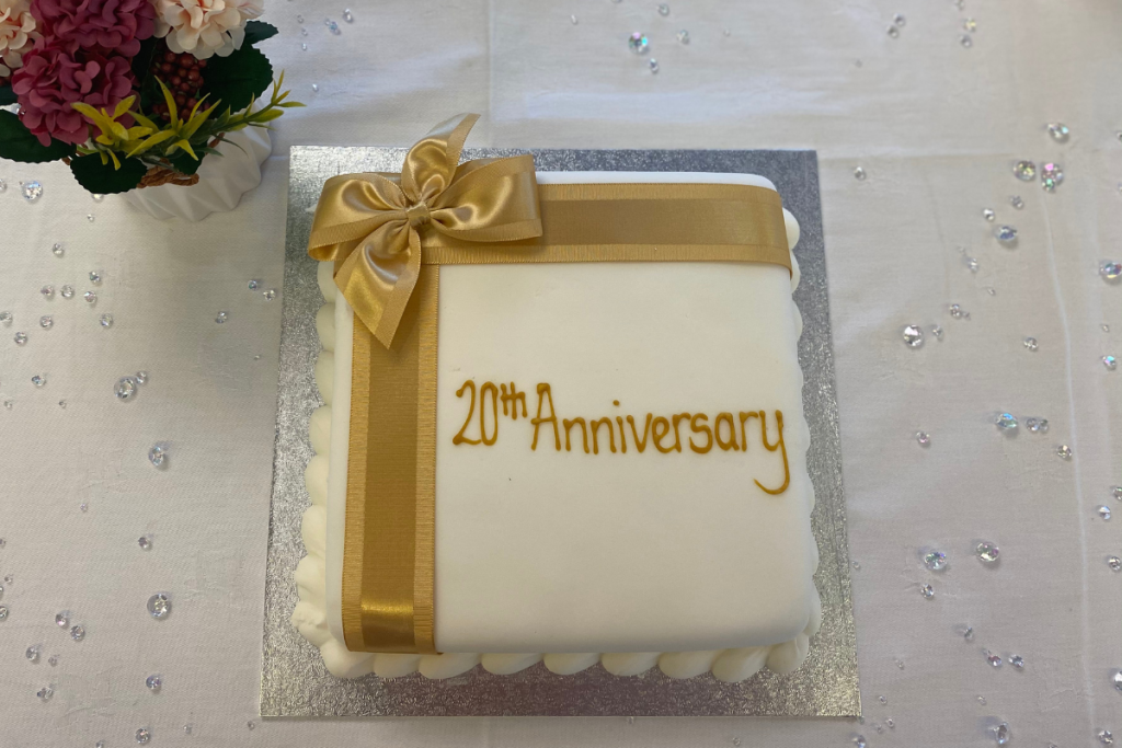A Square Cake, Covered In White Icing, With A Gold Bow. '20Th Anniversary' Is Written In Gold Icing.