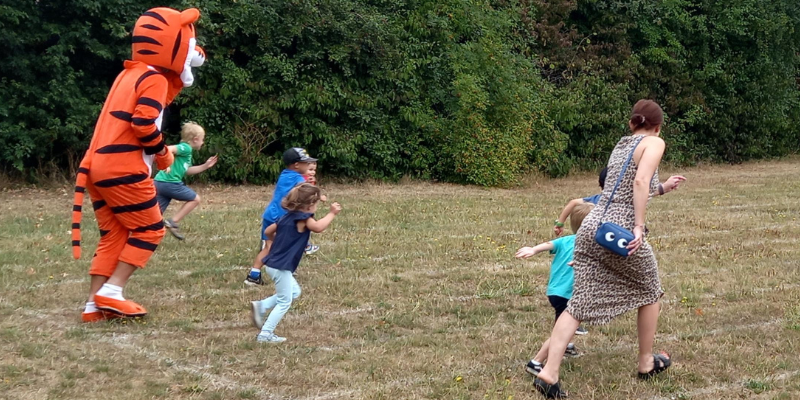 Children Are Seen Racing A Playworker Dressed As A Tiger On An Outdoor Running Track. Image From Last Year's Play In The Park