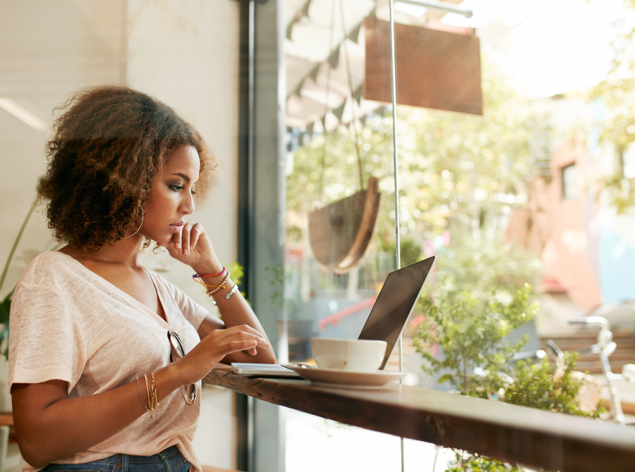 A Woman Sits At A Window Table In A Coffee Shop. She Is Working On Her Laptop With A Cappuccino Cup Beside Her