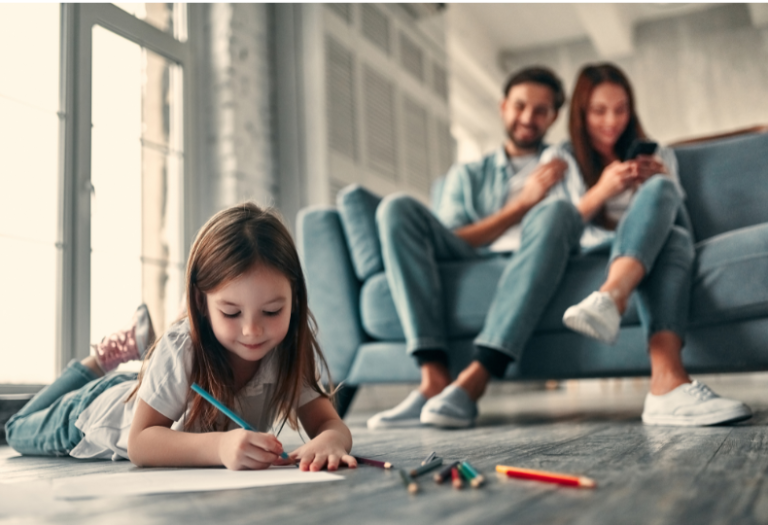 Little girl lying on the floor drawin,g with her parents looking on from the sofa
