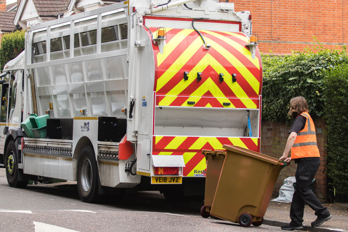 A Man In A High Vis Vest With Two Brown Wheely Bins Stands Next To A Refuse Truck