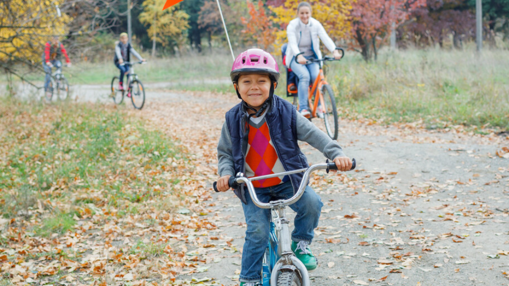A Boy On A Bike Looks Straight To Camera As He Rides On A Country Trail. Other Family Members Are On Bikes Just Behind Him