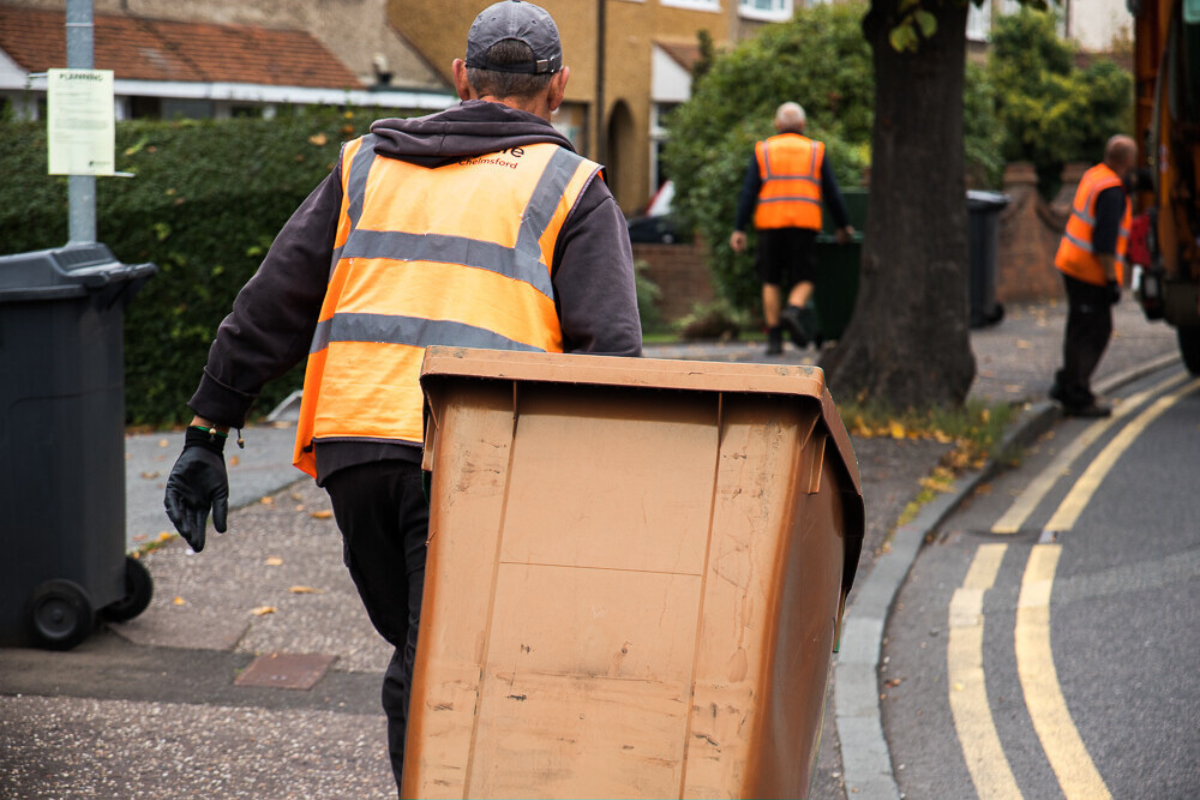 A Man In A High Vis Vest Pulls A Brown Wheely Bin Towards A Refuse Truck