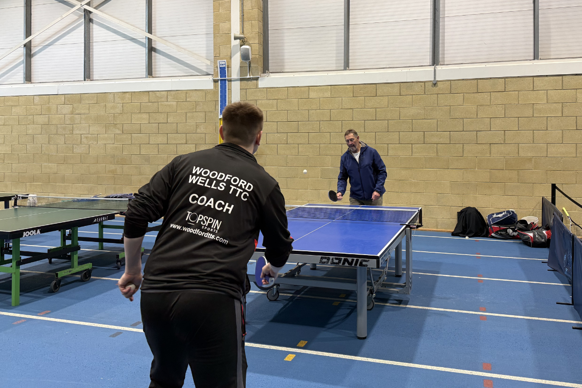 Graham Gooch playing table tennis with a volunteer while children break for lunch.png