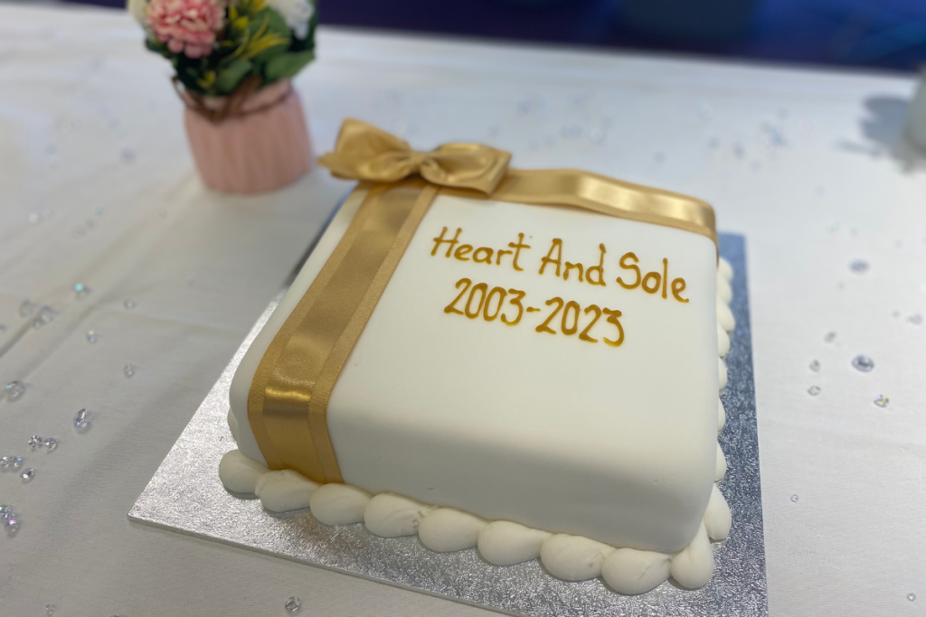 A Square Cake, Covered In White Icing, With A Gold Bow. The Words 'Heart And Sole 2003 2023' Are Written In Gold Icing.