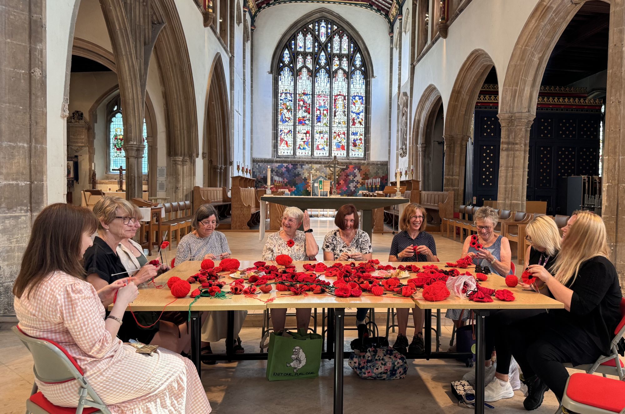 Ladies sitting around a table knitting poppies for Remembrance inside Chelmsford cathedral - poppy knit-a-thon landscape group photo