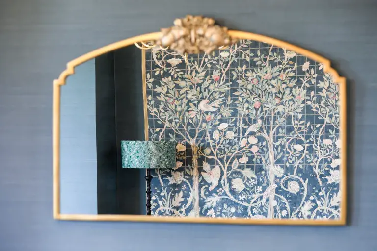 A Gold Mirror With A Reflection Of Some William Morris Wallpaper And A Lamp